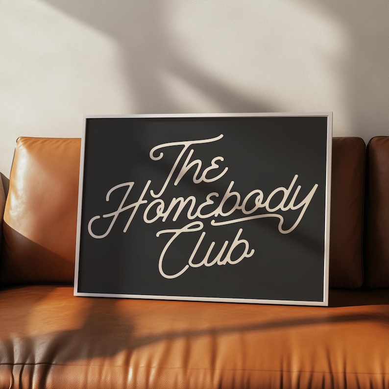 May include: A black framed print with white text that reads "The Homebody Club" in a cursive font.