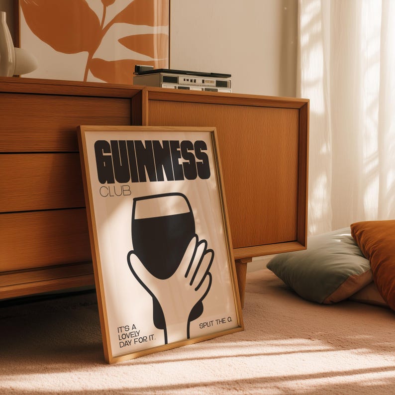 Pu&ograve; includere: Un poster incorniciato con le parole "GUINNESS CLUB" in grassetto nero. Il poster presenta una grafica nera di una mano che tiene una pinta di stout, con il testo "IT'S A LOVELY DAY FOR IT" e "SPLIT THE Q".