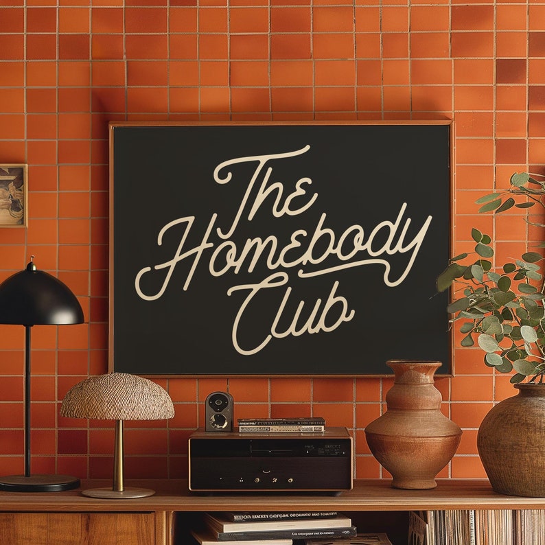 May include: A black framed sign with white text that reads "The Homebody Club" against a backdrop of orange tile.