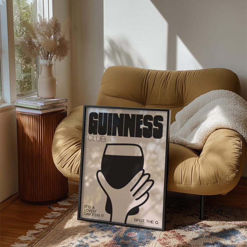 Pu&ograve; includere: Poster con cornice nera con la scritta "GUINNESS CLUB" in nero. Il poster presenta una grafica in bianco e nero di una mano che tiene una pinta di Guinness. Sono visibili anche i testi "IT'S A LOVELY DAY FOR IT" e "SPLIT THE Q". Il poster &egrave; appoggiato a una poltrona color senape.