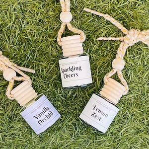 May include: Three glass car air fresheners with wooden tops and string hangers. The scents are Sparkling Cheers, Vanilla Orchid, and Lemon Zest.