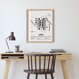 May include: A black and white print of the Dallas Fort Worth International Airport layout, framed in a light wood frame. The print is hanging on a white wall above a wooden desk with a black lamp and a chair.