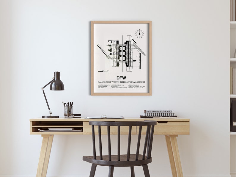 May include: A black and white print of the Dallas Fort Worth International Airport layout, framed in a light wood frame. The print is hanging on a white wall above a wooden desk with a black lamp and a chair.