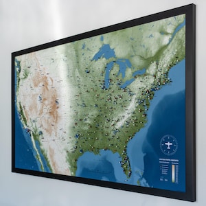 May include: A framed map of the United States, showcasing a detailed topographical view. The map highlights various airports with colored markers, set against a backdrop of green land and blue oceans. The frame is black, and the map includes a legend.