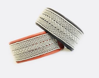 May include: Two leather cuff bracelets. One is black with silver-colored woven detailing, and the other is reddish-brown with similar silver-colored woven detailing. The bracelets are displayed on a white background.