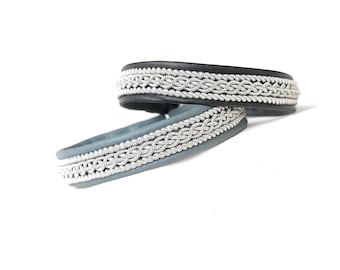 May include: Two leather bracelets, one black and one blue-grey, with intricate silver-colored braided detailing. The bracelets are displayed on a white background, showcasing their craftsmanship and design. The bracelets are a stylish accessory.
