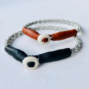 May include: Two braided bracelets with leather accents and white button closures. One bracelet is black and the other is brown.