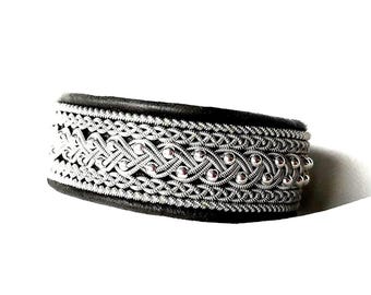 May include: A silver-toned bracelet with intricate woven metalwork and small, round, pink accents. The bracelet has a black backing and a detailed, textured design. The jewelry piece is set against a white background.
