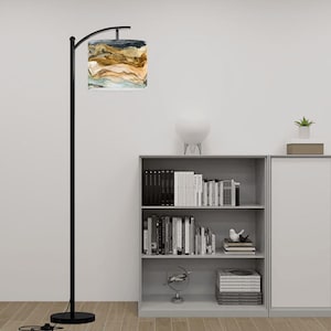 May include: A black arched floor lamp with a round base and a cylindrical lampshade. The lampshade features a watercolor design in shades of brown, beige, blue, and white. The lamp is positioned in front of a bookshelf.