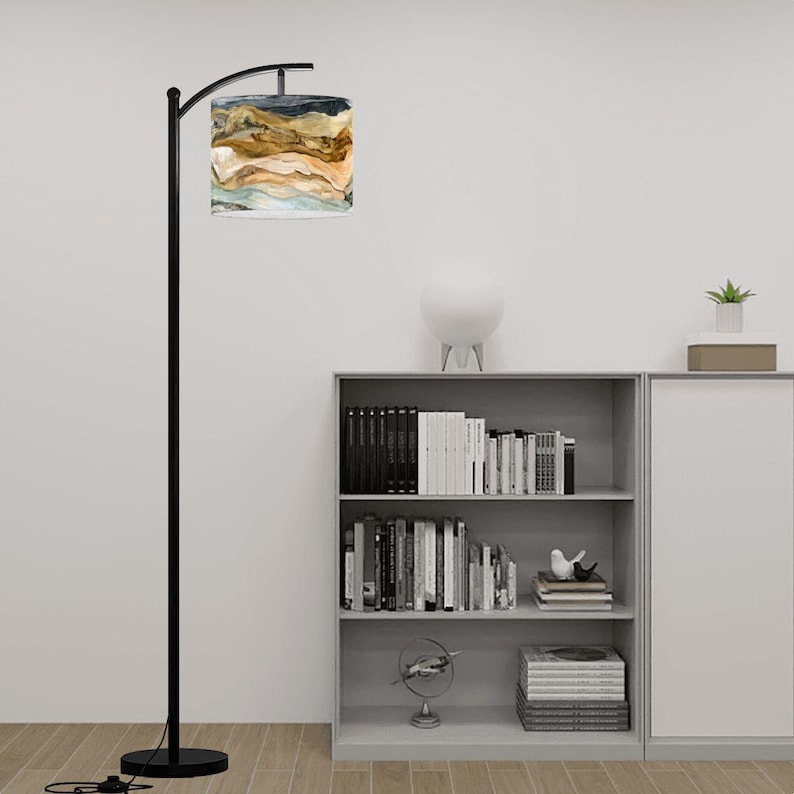 May include: A black arched floor lamp with a round base and a cylindrical lampshade. The lampshade features a watercolor design in shades of brown, beige, blue, and white. The lamp is positioned in front of a bookshelf.