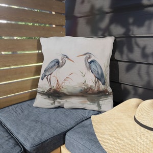 May include: A white throw pillow with a watercolor illustration of two blue herons standing in a marsh. The herons are facing each other and appear to be communicating. The background is a soft watercolor wash of blue and green.