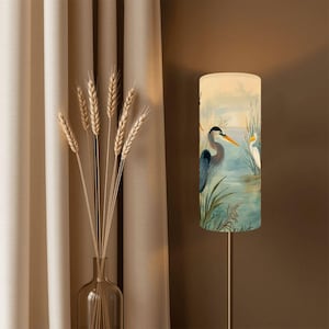 May include: A floor lamp with a cylindrical lampshade featuring a watercolor painting of two herons in a marsh setting. The lamp has a silver metal pole and is positioned near a beige curtain and a vase of wheat.