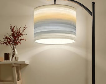May include: A modern floor lamp with a black metal frame and a cylindrical lampshade. The lampshade features horizontal bands of color in shades of yellow, blue, and gray. The lamp is turned on, illuminating the room.