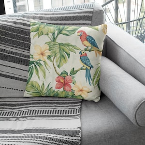 May include: A throw pillow with a tropical print featuring two parrots, green leaves, and pink and yellow flowers. The pillow is on a gray couch with a black and white striped throw blanket.