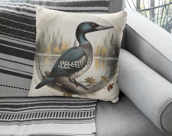 Può includere: Un cuscino con sfondo color crema che presenta un'illustrazione dettagliata di un loon in un ambiente boschivo. Il loon è nero, bianco e blu con un collo lungo e sottile e un occhio rosso. Lo sfondo è un'illustrazione in stile acquerello di alberi e un lago.