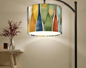 May include: A floor lamp with a black metal arched stand and a cylindrical drum shade. The shade features a repeating vertical pattern of abstract, elongated shapes in shades of yellow, orange, blue, green, and teal against a cream background.