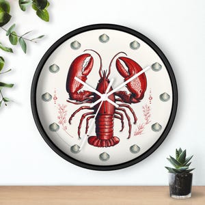 May include: A round wall clock with a black frame. The clock face features a red lobster illustration with white clock hands. Small seashell illustrations and red coral accents surround the lobster on a cream background.
