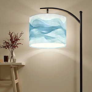 May include: A floor lamp with a black metal frame and a cylindrical lampshade. The lampshade features a light blue and white wavy pattern. A small table with a vase of red flowers and a candle sits to the left of the lamp.