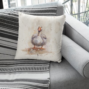 Peut inclure: Un coussin blanc avec une illustration aquarelle d'une oie grise debout sur une surface brun clair. L'oie a un bec orange et des pattes palmées.
