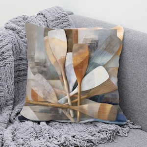 May include: Decorative square pillow featuring a watercolor-style design of overlapping wooden oars in shades of brown, beige, and blue. The pillow has a soft, textured appearance, suggesting a comfortable home decor item.