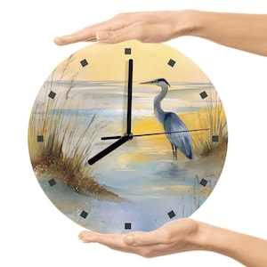 May include: Round wall clock featuring a watercolor painting of a blue heron standing in water. The clock face has a coastal scene with a yellow and blue sky, and tall grass. Black clock hands and square hour markers.