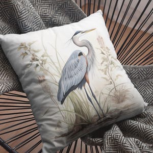 May include: A decorative pillow with a watercolor illustration of a great blue heron in a marsh setting. The heron is depicted with blue and gray feathers, standing among reeds and grasses. The pillow is on a black chair with a patterned blanket.