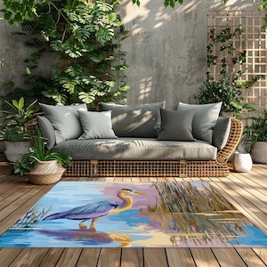 May include: Outdoor rug featuring a heron standing in water, with a reflection. The rug has a blue, yellow, and purple color palette. A gray sofa with pillows and plants are in the background. The rug is placed on a wooden deck.