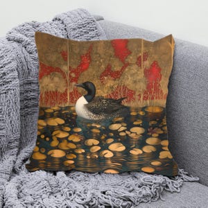 May include: Square cushion with a detailed illustration of a loon on a pond with lily pads. The background has a stylized design with red and gold tones. The cushion is resting on a grey blanket.
