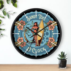 May include: Round wall clock with a black frame. The clock face features a tropical scene with a woman, surfboard, palm trees, and flowers. Text on the clock face reads "Sandy Toes O'Clock". The clock has white hour markers.