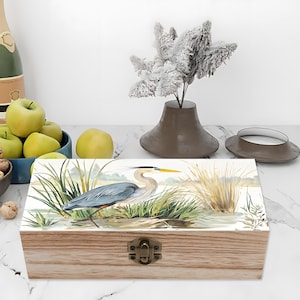 May include: Wooden box with a hinged lid and a metal clasp. The lid features a watercolor painting of a blue heron standing in water with tall grass. The box is made of light-colored wood.