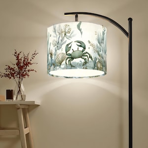 May include: A floor lamp with a cylindrical lampshade featuring a sea-themed design. The lampshade showcases a light blue crab, seashells, and aquatic plants against a white background. The lamp has a black, curved arm and base.