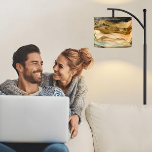 May include: A black floor lamp with a lampshade featuring an abstract landscape design in shades of brown, beige, and teal. A couple is smiling while looking at a laptop on a white couch.