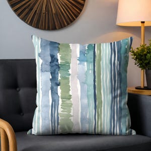 May include: Square cushion featuring a watercolour stripe design in shades of blue, green, and white. The cushion is on a dark grey sofa. A wooden sunburst wall decor is visible in the background.