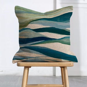 May include: A square throw pillow featuring an abstract landscape pattern with layered horizontal bands of teal, dark blue, sage green, and cream. The fabric has a visible woven texture, and the design mimics rolling hills or waves.