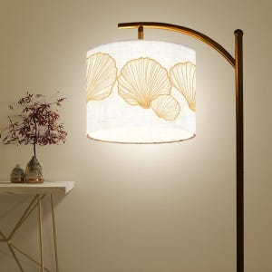 May include: A floor lamp with a curved, bronze-colored metal frame. The lampshade is cylindrical, white, and features a repeating pattern of gold-colored, stylized leaf designs. The lamp is turned on, illuminating the room.