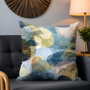 May include: Square decorative pillow with an abstract design. The pillow features a pattern of overlapping shapes in shades of blue, yellow, and brown. The design has a watercolor effect, with soft edges and subtle textures. The pillow is placed on a dark gray couch.