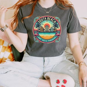 May include: A gray t-shirt with a vintage-style graphic of the South Beach skyline. The graphic features palm trees, a beach, and the text "South Beach Art Deco Paradise 1970-1920".