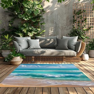 May include: A rectangular outdoor rug featuring a beach scene with turquoise water, white waves, and a sandy shore. The rug is placed on a wooden deck, with a wicker sofa and gray cushions in the background, surrounded by plants.