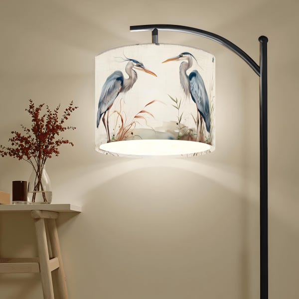 Może przedstawiać: Czarna metalowa lampa podłogowa z cylindrycznym kloszem. Klosz przedstawia akwarelowy wzór dwóch niebieskich czapli w krajobrazie bagiennym. W pobliżu znajduje się mały stolik z wazonem z czerwonymi kwiatami.
