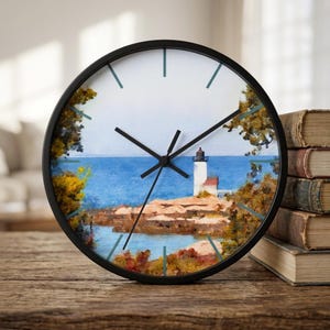 May include: A round wall clock with a black frame and a white face featuring a painted coastal scene. The image depicts a white lighthouse on a rocky shore surrounded by autumn-colored trees, overlooking a blue ocean under a pale sky.