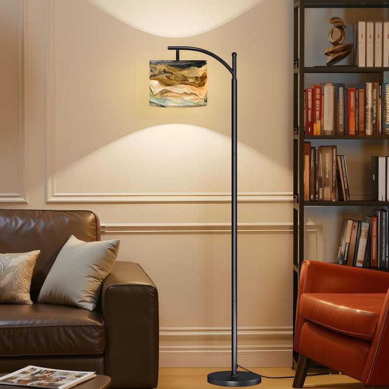 May include: A black arc floor lamp with a rectangular lampshade featuring a landscape design in shades of brown, beige, and blue. The lamp is positioned next to a brown leather sofa and a bookshelf.