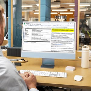 Op de afbeelding: Een desktopcomputer met een spreadsheet met de titel "Tip Tracker" op een houten bureau. Een wit toetsenbord, muis en notitieblok staan voor de monitor. Een witte waterfles staat rechts van de monitor.