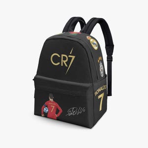 May include: A black backpack featuring the gold CR7 logo on the front, a graphic of a soccer player in a red jersey with the number 7, a white signature, and the name Ronaldo with the number 7 on the side, along with various soccer team logos.