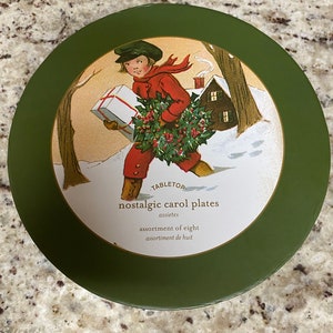 Puede incluir: Lata redonda verde con una ilustración vintage de un niño que lleva una corona y una caja de regalo. El texto en la lata dice "Tabletop nostalgic carol plates, assiettes, assortment of eight, assortment de huit."
