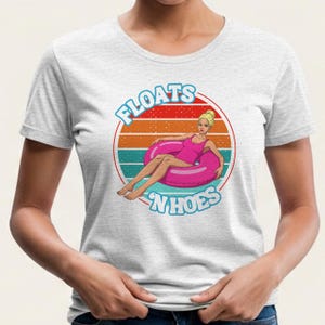 May include: A light gray t-shirt featuring a retro design with the words "FLOATS 'N HOES" in blue and a woman in a pink swimsuit lounging on a pink inflatable ring. The background has a sunset design.