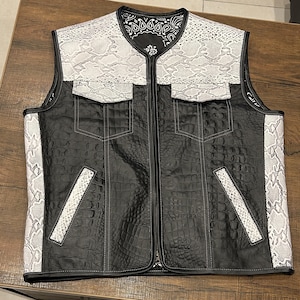 Könnte beinhalten: Eine schwarze Lederweste mit weißen Schlangenleder-Akzenten. Die Weste hat einen Reißverschluss und zwei große Taschen auf der Vorderseite. Die Weste ist mit einem schwarz-weißen Bandana-Muster gefüttert.