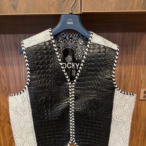 Pode incluir: Colete de couro preto com acabamento em pele de cobra branca e um design branco costurado. O colete tem um fecho de correr e uma etiqueta que diz "ROCKY".