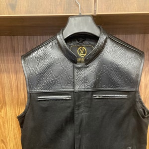 May include: Black leather vest with a snakeskin pattern on the front and back. The vest has a zipper closure and two pockets on the front. The vest has the text "THE V" on the front.
