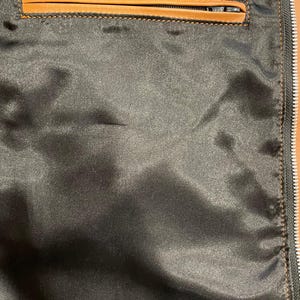 Puede incluir: Primer plano del forro de una chaqueta de sat&eacute;n negro con un bolsillo ribeteado en cuero marr&oacute;n. El bolsillo tiene cierre de cremallera. La cremallera de la chaqueta es visible en el lado derecho.