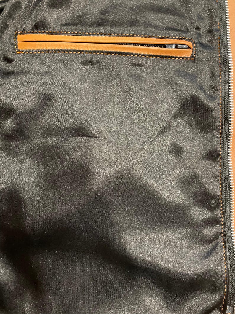 Puede incluir: Primer plano del forro de una chaqueta de sat&eacute;n negro con un bolsillo ribeteado en cuero marr&oacute;n. El bolsillo tiene cierre de cremallera. La cremallera de la chaqueta es visible en el lado derecho.